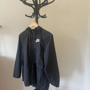 Svart Nike vindjacka med huva - Svart vindjacka från Nike med huva och dragkedja framtill. Jackan har en klassisk sportig look, justerbar nederkant med snörning och meshfoder på insidan för extra komfort. Perfekt för blåsiga dagar och streetstyle. Nike-logga på bröstet.