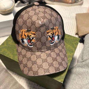 Gucci keps med tigertryck och GG-mönster - Snygg keps från Gucci med klassiskt GG-mönster i beige och brunt, två stora tigertryck framtill och svart mesh på sidorna och bak. Justerbar rem med grönt och rött band bak. Perfekt statement-piece för dig som vill sticka ut. Låda ingår inte
