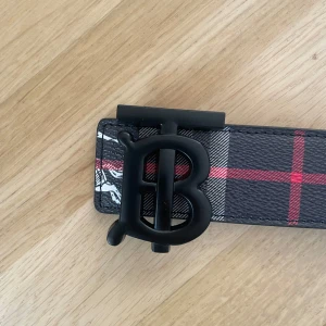 Svart burberry bälte - Snyggt bälte med rutigt mönster i svart, grått, vitt och rött. Bältet har ett stort matt svart B-format spänne och är dekorerat med vita och röda detaljer längs hela bandet. Perfekt för att lyfta vilken outfit som helst.