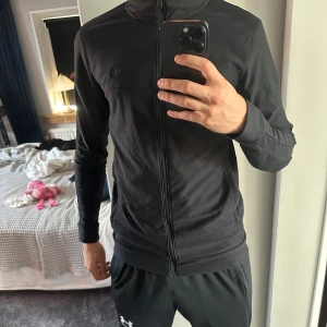 Svart ziptröja från Under Armour - Svart långärmad ziptröja från Under Armour med diskret logga på bröstet. Tröjan har en hel dragkedja framtill och är gjord i ett mjukt syntetmaterial som andas. Perfekt för träning eller chill, med en clean och sportig look. Fickorna har dragkedjor och det finns även innerfickor.
