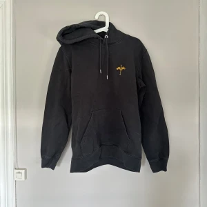 Frej Larsson merch - Svart hoodie med huva och snörning, samt en gul broderad detalj på bröstet. Frej Larsson merch