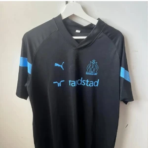 Svart Marseille fotbollströja Puma - Snygg svart fotbollströja från Olympique de Marseille med ljusblå detaljer och logga på bröstet. Puma-loggan och sponsortryck i blått framtill, samt blå prickiga ränder på ärmarna. Tröjan har en modern passform och är tillverkad i lätt polyester.