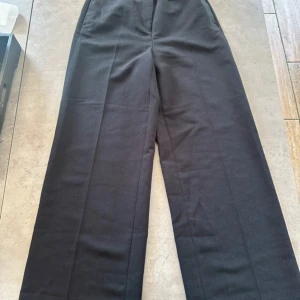 Svarta kostymbyxor från H&M - Snygga svarta kostymbyxor från H&M med klassisk rak passform och pressveck. Byxorna har hög midja och två bakfickor med knapp. Perfekta för en clean och stilren look. Materialet är mjukt och känns bekvämt mot huden.