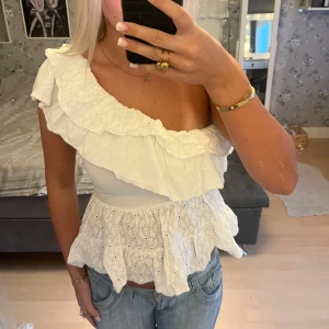 Vit omlott blus med volang - Superfin vit blus med ett axelband och volangdetaljer runt halsringningen och nertill. Toppen har broderade spetsdetaljer och är omlott med en luftig, somrig känsla. Perfekt till jeans eller kjol för en fräsch look.