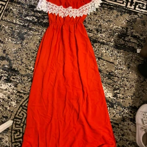 Röd långklänning med spetsdetalj - Snygg röd långklänning med offshoulder-modell och vit spets längs överdelen. Klänningen har resår i midjan för en bekväm passform och ett luftigt fall. Perfekt för dig som vill sticka ut med en färgstark look.