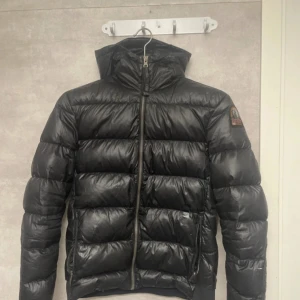 Svart dunjacka Parajumpers Sheen YL - Snygg svart dunjacka från Parajumpers, modell Sheen. Jackan har en glansig finish, huva och klassisk patch på ärmen. Fylld med dun för extra värme och har dragkedja framtill samt två fickor med dragkedja. Perfekt för kalla dagar och riktigt street. Plus den har lite håll i fickorna 