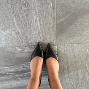 Svarta pumps med spetsig tå och rem - Snygga svarta pumps med spetsig tå och smal klack. Skorna har en elegant rem runt hälen med spänne och är tillverkade i ett slätt material som ger en stilren look. Perfekta för dig som gillar klassisk och trendig stil.