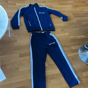 Blåa träningsbyxor Palm Angels - Snygga blåa träningsbyxor från Palm Angels med vita sidoränder och broderad logga på benet. Byxorna har elastisk midja och raka ben, perfekta för en sportig och trendig look. Materialet är mjukt och följsamt, passar till streetwear.