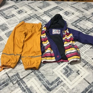 Randig regnjacka från Lupilu, 110/116 - Färgglad regnjacka från Lupilu i storlek 110/116. Jackan har lila ärmar, huva och insida, samt randigt mönster i gult, rosa, vitt och lila. Stängs med dragkedja och har reflexdetaljer. Perfekt för regniga dagar och lek utomhus.