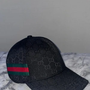 Svart keps med GG-mönster - Inte använd eftersom att jag fick den på min födelsedag samtidigt som jag fick en sån av min vän på samma dag så jag hade ingen nytta av den, bara använd till att ta bilder med inget annat. Snygg svart keps från Gucci med klassiskt GG-monogram över hela kepsen. På sidan finns en grön och röd rand som ger den en lyxig touch. Justerbar rem baktill i skinn och insida med Gucci-logga. Perfekt accessoar för att lyfta din outfit.