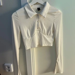 Vit croppad ribbad polotopp från Shein - Säljer en vit croppad polotopp från Shein med långa ärmar och ribbad struktur. Toppen har krage, knappar framtill och är figurnära. Perfekt att styla med jeans eller kjol för en trendig look.
