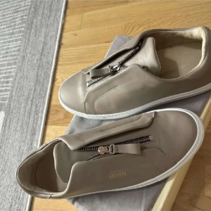 Beige sneakers från Axel Arigato - Snygga beige sneakers från Axel Arigato i skinn med vit sula och dragkedja på ovansidan istället för klassisk snörning. Minimalistisk design med diskret guldfärgad logga på sidan. Perfekta för dig som gillar stilrena och moderna skor.