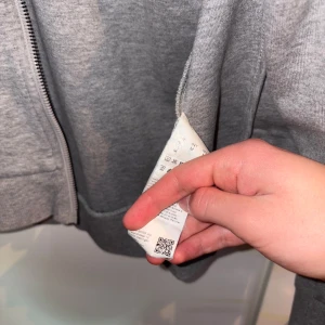 Grå zip-hoodie från BOSS - Grå hoodie med dragkedja från BOSS Hugo Boss. Jackan har huva och röd broderad logga på bröstet. Perfekt för en avslappnad stil och enkel att matcha med andra plagg. OG Hugo Boss