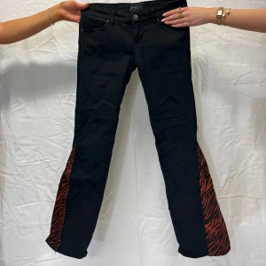 Svarta jeans med röd zebra - Svarta lågmidjade jeans med röd zebra. Storlek: 36. Mått: Innerbenslängd: 80 cm, Midjemått: 80 cm.