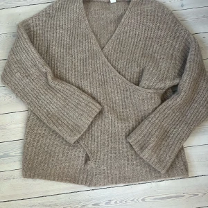 Beige stickad omlottkofta från Kay/day - Mysig beige stickad omlottkofta från Kay/day x Sanna Jörnlind Benman. Tröjan har v-ringning och långa ärmar, perfekt för lager på lager. Snygg ribbad struktur och avslappnad passform.