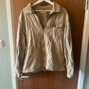 Beige jacka med dragkedja från East West - Säljer en beige jacka från East West i storlek M. Jackan har dragkedja framtill, en bröstficka med knapp och klassisk krage. Perfekt för vår och höst, enkel och stilren design.