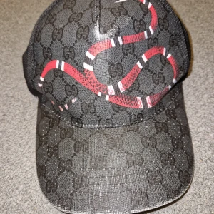 Svart keps med ormtryck  - Säljer en svart keps från med klassiskt GG-mönster och en röd orm som slingrar sig över framsidan. Kepsen har böjd skärm och justerbar rem baktill med blå-röd detalj. Tillverkad i Italien.