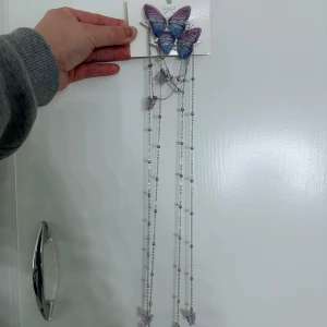 Silverfärgat halsband med lila fjärilar - Långt halsband i silverfärgad metall med flera hängen i form av lila fjärilar. Kedjan har små silverkulor och fjärilarna är i olika storlekar och nyanser av lila. Perfekt för dig som gillar unika och lekfulla accessoarer med naturtema.