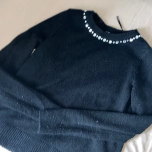 Svart stickad tröja med pärldetaljer - Säljer en svart stickad tröja med långa ärmar och rund halsringning. Tröjan har snygga pärldetaljer längs halsen som ger en extra touch. Perfekt att styla till olika outfits!