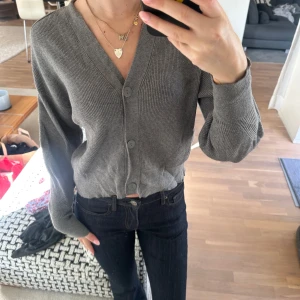 Grå ribbad kofta med knappar - Säljer en grå croppad kofta med v-ringning och stora knappar framtill. Tröjan är ribbstickad och har långa ärmar.