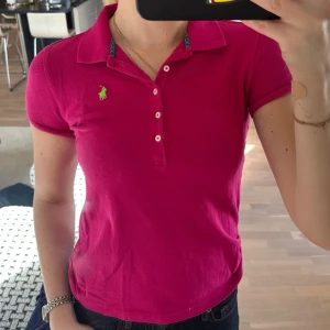 Ceriserosa pikétröja från Ralph Lauren - Säljer en ceriserosa pikétröja från Ralph Lauren med klassisk krage och knappslå framtill. Tröjan har korta ärmar och grönt broderat logotyp på bröstet samt en grön detalj i sidan nertill. Den har ett litet hål vid sidan men om man kollar på bild tre är det inget jätte synligt