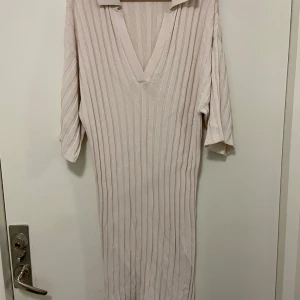 Ljusbeige ribbad långklänning med krage - Klänningen är helt ny och oanvänd! Säljer en ljusbeige ribbad långklänning med v-ringning och krage. Klänningen har trekvartsärm och slits på sidorna för extra rörelsefrihet. Perfekt för dig som gillar en stilren och avslappnad look.