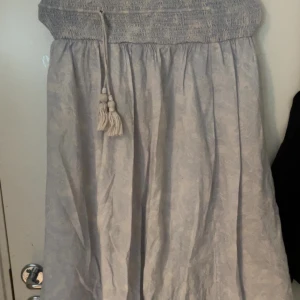 Ljusblå långkjol med knytband från Gina Tricot - Säljer en ljusblå långkjol från Gina Tricot med diskret mönster och bred smock i midjan. Kjolen har dekorativa knytband med tofsar framtill och ett lätt, luftigt tyg som passar perfekt till varmare dagar.
