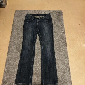 Mörkblå jeansbyxor som är utsvängda med detaljer på. - Mörkblå jeansbyxor med vita kontrastsömmar och snygga detaljer på bakfickorna, bland annat dragkedja. Klassisk femficksmodell med bootcut passform och låg midja. Storleken 36/38 