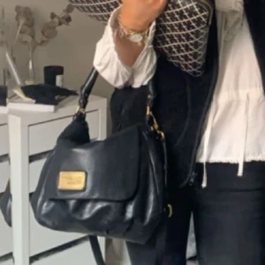 Svart axelväska från Marc by Marc Jacobs - Snygg svart axelväska i skinn från Marc by Marc Jacobs med guldiga metalldetaljer och logoplatta framtill. Väskan har justerbar axelrem och lockstängning. (Lite defekter i 2 innerfickor, skriv för fler bilder på det)