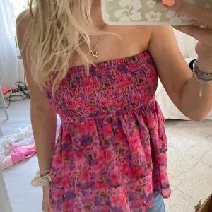 Blommig tubtopp i rosa och lila - Säljer en söt blommig tubtopp i rosa och lila nyanser. Toppen har smock upptill och volangig nederdel, perfekt för varma dagar. Mjukt och lätt material som sitter bekvämt. Passar alla upp till M då den är stretchig