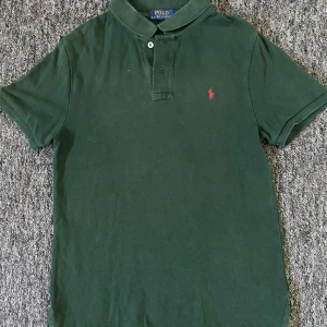 Grön pikétröja från Polo Ralph Lauren - Säljer en mörkgrön pikétröja från Polo Ralph Lauren i slim fit-modell. Tröjan har korta ärmar, klassisk krage med två knappar och en liten röd broderad logga på bröstet. Perfekt för en clean och stilren look.