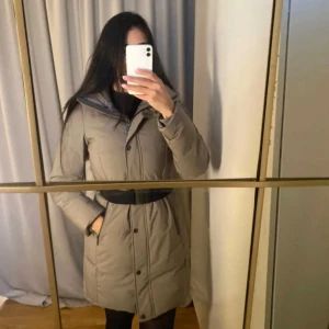 Beige dunjacka med bälte - Säljer en beige grön lång dunjacka med bälte i midjan och knappar framtill. Jackan har hög krage och långa ärmar, perfekt för kyliga dagar. Snygg och stilren modell som passar till många tillfällen.