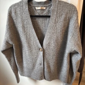Grå stickad kofta - Mysig grå stickad kofta med v-ringning och en knapp framtill. Koftan har lång ärm och ribbade muddar vid ärmslut och nederkant. Perfekt att slänga på sig när det är kyligt ute.