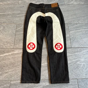 Evisu jeans - Säljer ett par svarta EVISU jeans med vit målad båge och röda blommor på baksidan. Klassisk femficksmodell med raka ben och läderpatch i midjan. Rå denim med tydliga kontrastsömmar och unik design. DM för mer info.