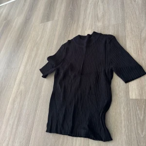 Svart ribbad topp med halvärm - Säljer en svart ribbad topp med hög hals och halvärm. Toppen har en tight passform och är perfekt att styla till olika outfits. Materialet är stretchigt och bekvämt.