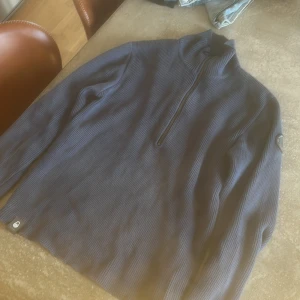 Blå stickad tröja med halv dragkedja från Sail Racing - Säljer en blå stickad tröja från Sail Racing med halv dragkedja framtill och hög krage. Tröjan har ribbad struktur och Sail Racing-logga på ärmen. Perfekt för kyliga dagar.