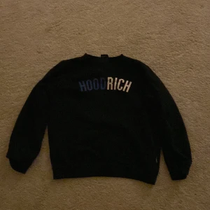 Svart sweatshirt från Hoodrich - Svart sweatshirt från Hoodrich med tryckt logga i vitt och blått på bröstet. Klassisk rund hals och långa ärmar. Perfekt för en avslappnad streetwear-look.