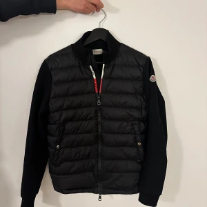Svart dunjacka från Moncler - Snygg svart dunjacka från Moncler med quiltad framsida och släta svarta ärmar. Jackan har dragkedja framtill, ståkrage och Moncler-logga på ena ärmen. Perfekt för dig som vill ha en stilren och sportig look.