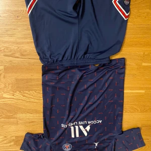 PSG fotbollströja och shorts - Mörkblå PSG fotbollströja med små röda detaljer och vita loggor, samt matchande shorts. Tröjan har korta ärmar och klubbmärke på bröstet. Materialet är lätt och andas, perfekt för träning eller match.