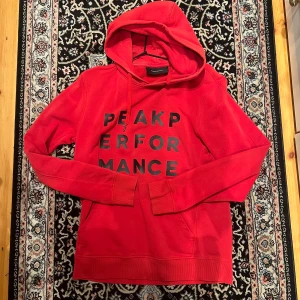 Röd hoodie från Peak Performance - Säljer en röd hoodie från Peak Performance med svart tryck på bröstet. Tröjan har huva med dragsko och en klassisk känguruficka framtill. Perfekt för en avslappnad stil.