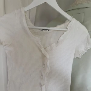 Vit ribbad topp med volangkant från Monki - Säljer en söt vit ribbad topp från Monki i storlek XS. Toppen har korta ärmar, knappar framtill och volangkant längs knäppningen och halsringningen. 