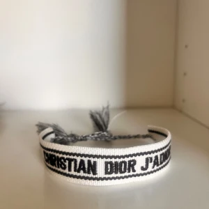 Vitt armband från Christian Dior - Snyggt vitt armband från Christian Dior med svart text 'CHRISTIAN DIOR J'ADIOR'. Armbandet har justerbar knytning med tofsar i ändarna och är perfekt som accessoar för att lyfta din outfit. Vet ej om de e äkta men tror det
