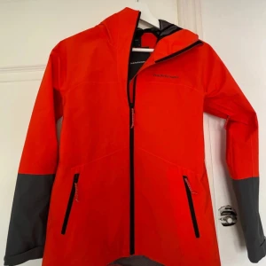 Orange vindjacka från Peak Performance - Snygg orange vindjacka från Peak Performance med svarta detaljer och gråa ärmslut. Jackan har dragkedja framtill och två fickor med dragkedja. Perfekt för blåsiga dagar och outdoor-aktiviteter.