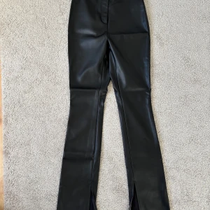 Svarta skinnbyxor med slits från NA-KD - Säljer ett par svarta skinnbyxor från NA-KD i storlek 34. Byxorna har en tight passform upptill och vida ben med slits framtill. Snyggt midjehög modell som ger en trendig look. Perfekta till sneakers eller klackar.