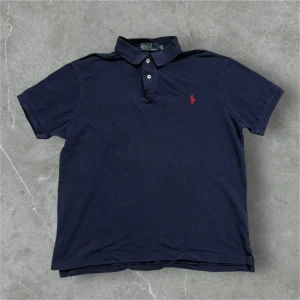 Ralph Lauren piké mörkblå  - Snygg mörkblå pikétröja från Ralph Lauren med klassisk krage och knappar. Den har en röd broderad logotyp på bröstet. Perfekt för en stilren look.