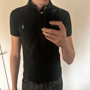 Svart pikétröja från Ralph Lauren - Säljer en svart pikétröja från Ralph Lauren med klassisk krage och korta ärmar. Tröjan har den ikoniska blå loggan broderad på bröstet och knäppning vid halsen. Perfekt för en stilren look. Storlek s slimfit . Toppen skick 
