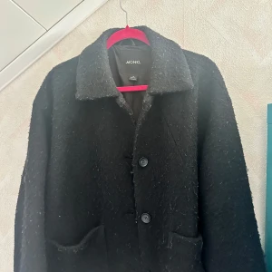 Svart kappa från Monki - Säljer en svart kappa från Monki med klassisk krage och två stora knappar framtill. Jackan har två fickor och en rak, lite oversized passform. Perfekt för kyligare dagar och enkel att matcha med olika outfits. Lite nopprig runt jackan. 