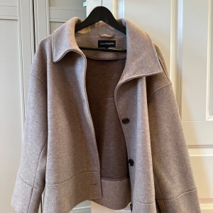 Beige kappa från KappAhl - Stilren beige kappa från KappAhl med klassisk krage och stora svarta knappar framtill. Jackan har lång ärm och rak passform, tillverkad i ett mjukt ullblandat material. Perfekt för kyligare dagar.