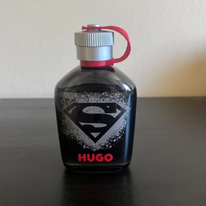 Hugo Boss parfym Superman Edition - Cool svart parfymflaska från Hugo Boss med Superman-logga på framsidan och röd text. Flaskan har en silverfärgad kork med röd snodd och rymmer 125 ml. Perfekt för dig som gillar unika samlarutgåvor och snygg design. använd par tre gånger pris diskuteras.