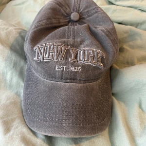 Grå keps med broderad text 'NEW YORK' - Snygg grå keps med broderad text 'NEW YORK EST. 1625' framtill och 'NEW YORK CITY' baktill. Justerbar rem baktill för perfekt passform. Perfekt accessoar för en avslappnad stil.